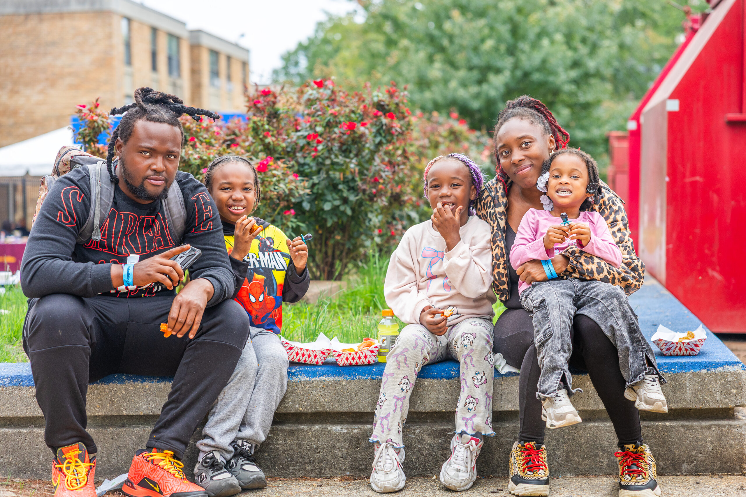 A DCHA family with three young children sits together on a curb and enjoy food.